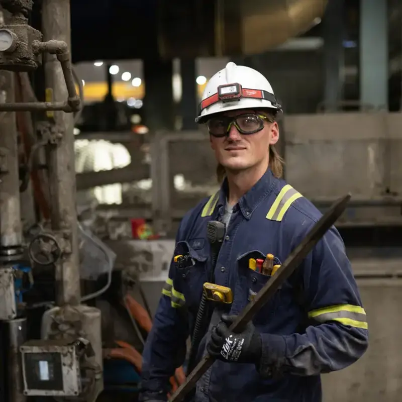 Portrait of a North Star BlueScope Steel worker in a blue uniform, safety glasses, and a white hard hat with a headlamp, holding a metal industrial tool.