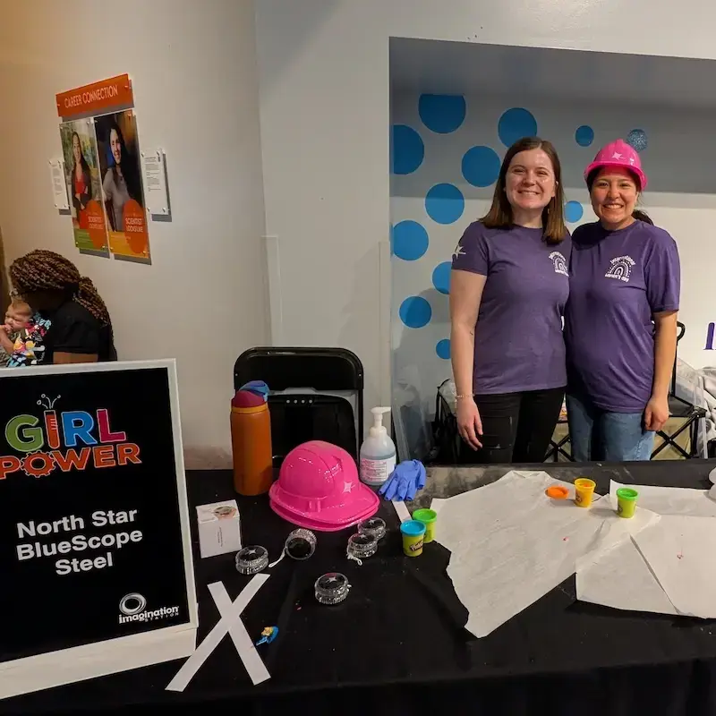 wo women in purple International Women's Day shirts at a "Girl Power" event booth for North Star BlueScope Steel, featuring pink hard hats and STEM activities.