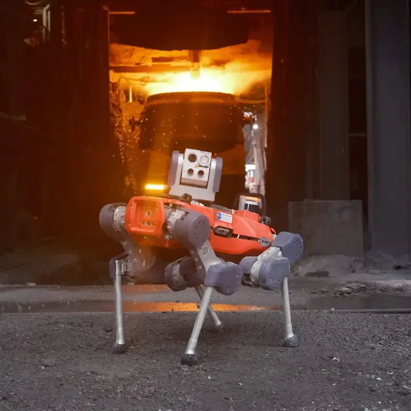 A sophisticated four-legged orange robotic dog stands on a gravel floor in front of a glowing industrial furnace at a steel manufacturing plant.